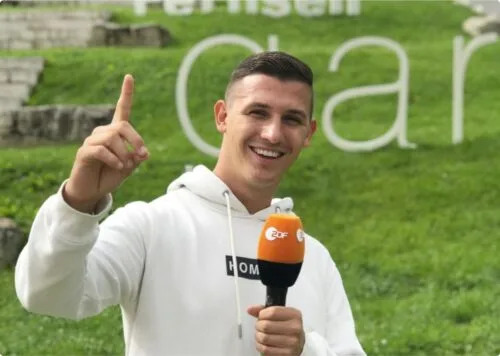 André Ortolf segurando um microfone e sorrindo, posando para a câmera em um ambiente ao ar livre.