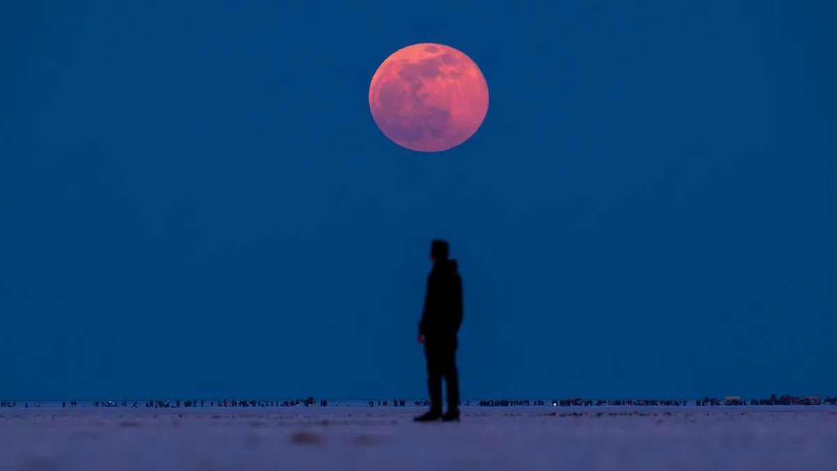 Pessoa observa uma lua de sangue no horizonte, com coloração vermelha intensa em contraste com o céu azul-escuro.