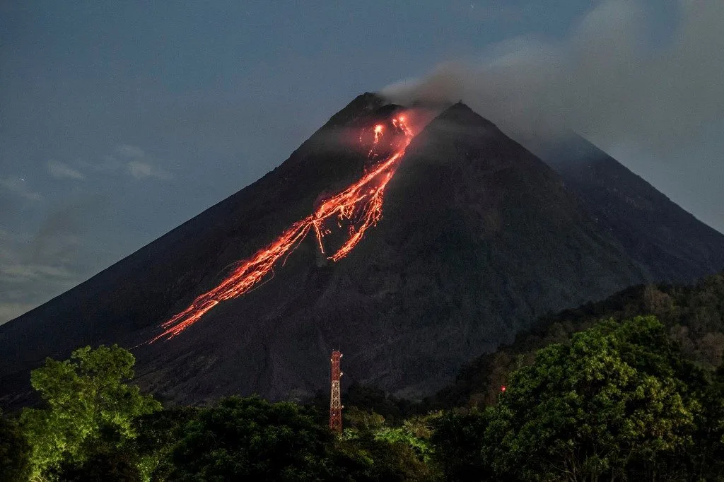 Erupção do vulcão Merapi lançando fumaça e lava em meio à vegetação tropical.